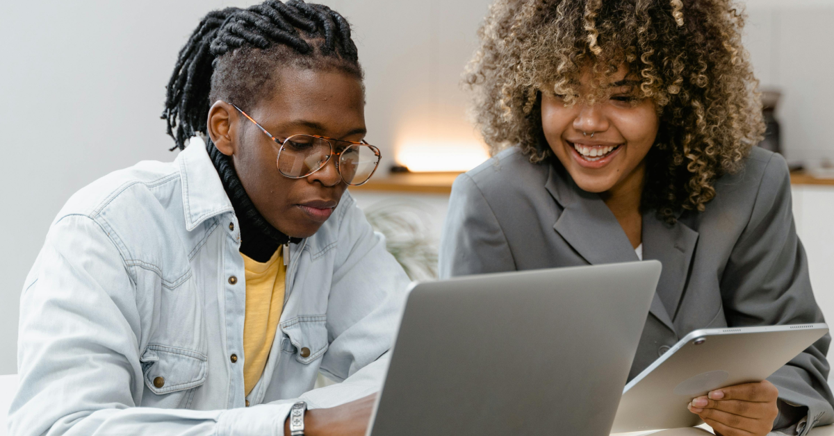 A imagem mostra dois jovens sentados lado a lado em um escritório. A esquerda, um homem negro de cabelos longos de dread e presos. Ele usa óculos de grau, camisa amarela, jaqueta jeans e olha para a tela de um computador. A direita, uma mulher negra de cabelos cacheados e soltos. Ela usa um paletó cinza e sorri, olhando para a mesma tela de computador que o seu colega de trabalho.